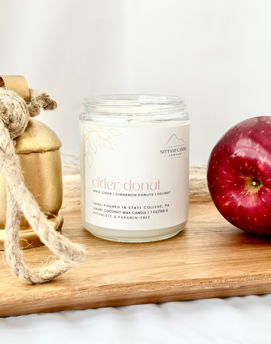 A glass jar candle labeled 'cider donut' by Nittany Candle Co., styled on a wooden board with a red apple and a gold bell prop against a white background.