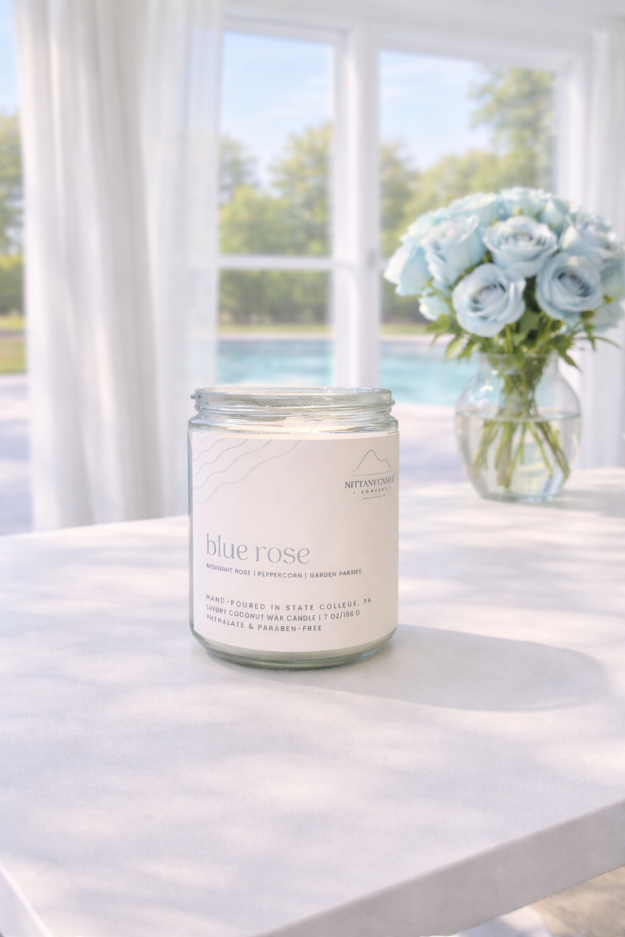 Candle labeled 'Blue Rose' on a table with a view of water and trees.