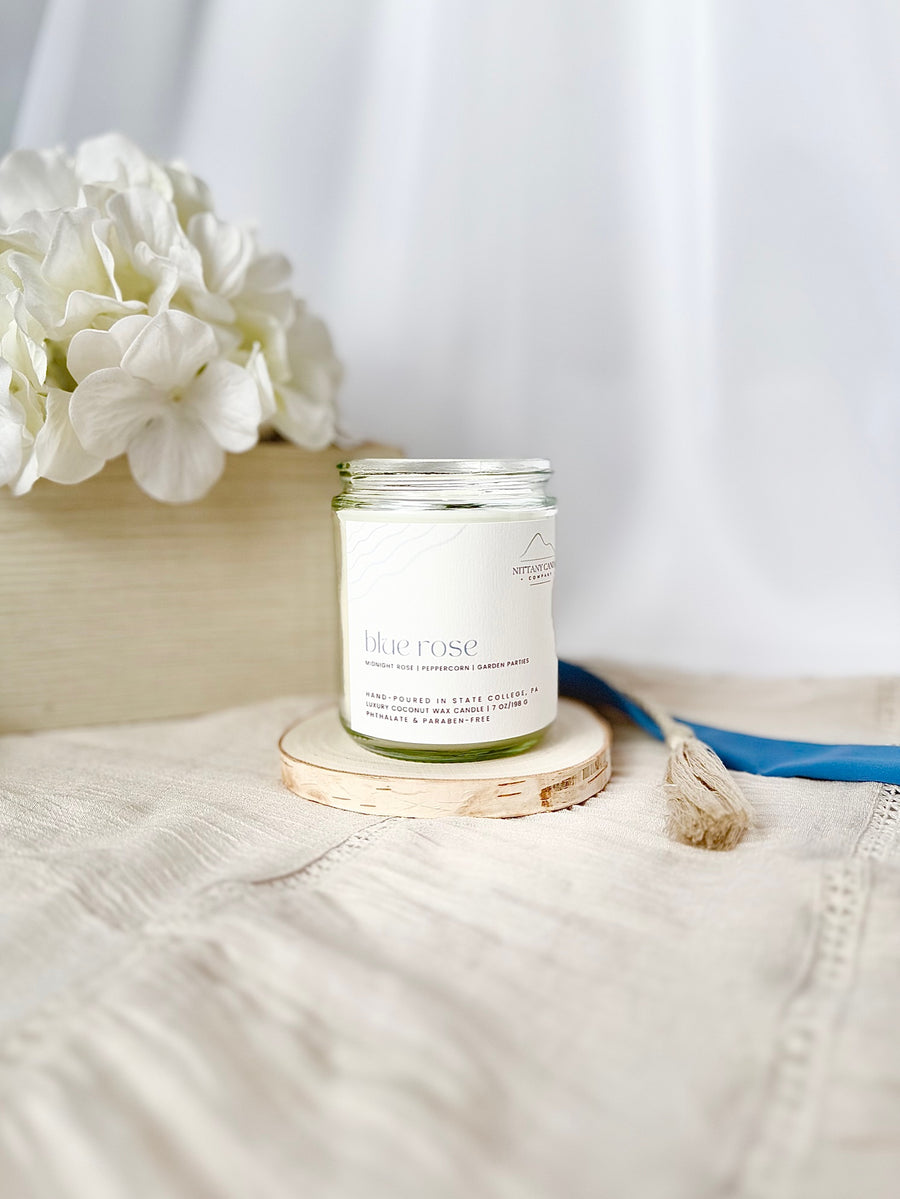 Candle labeled 'Blue Rose' on a wooden coaster with white flowers in the background