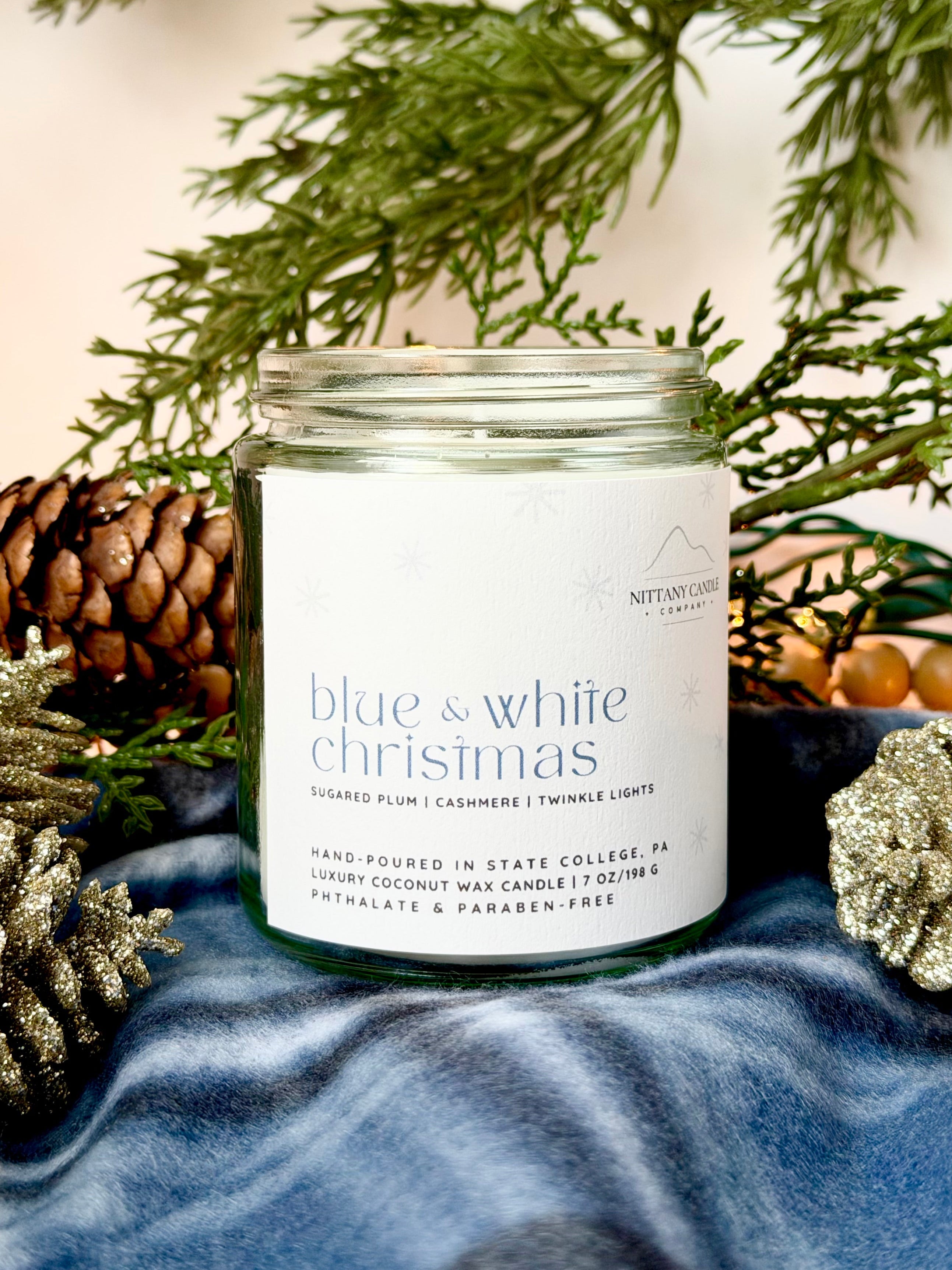 Penn State themed candle jar labeled 'Blue & White Christmas' surrounded by pine cones and branches on a textured surface.
