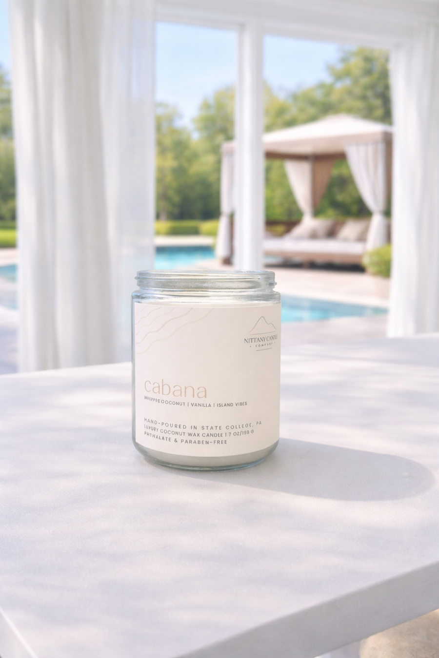A coconut scented candle labeled "cabana" on a white table with a pool and cabana in the background