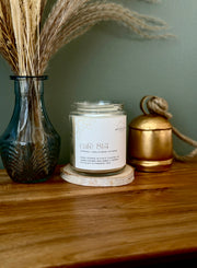 A glass jar candle labeled 'cafe 814' sits on a round wooden coaster on a wooden table, styled with a blue glass vase of dried grass on the left and a gold metal container on the right against a muted green wall.