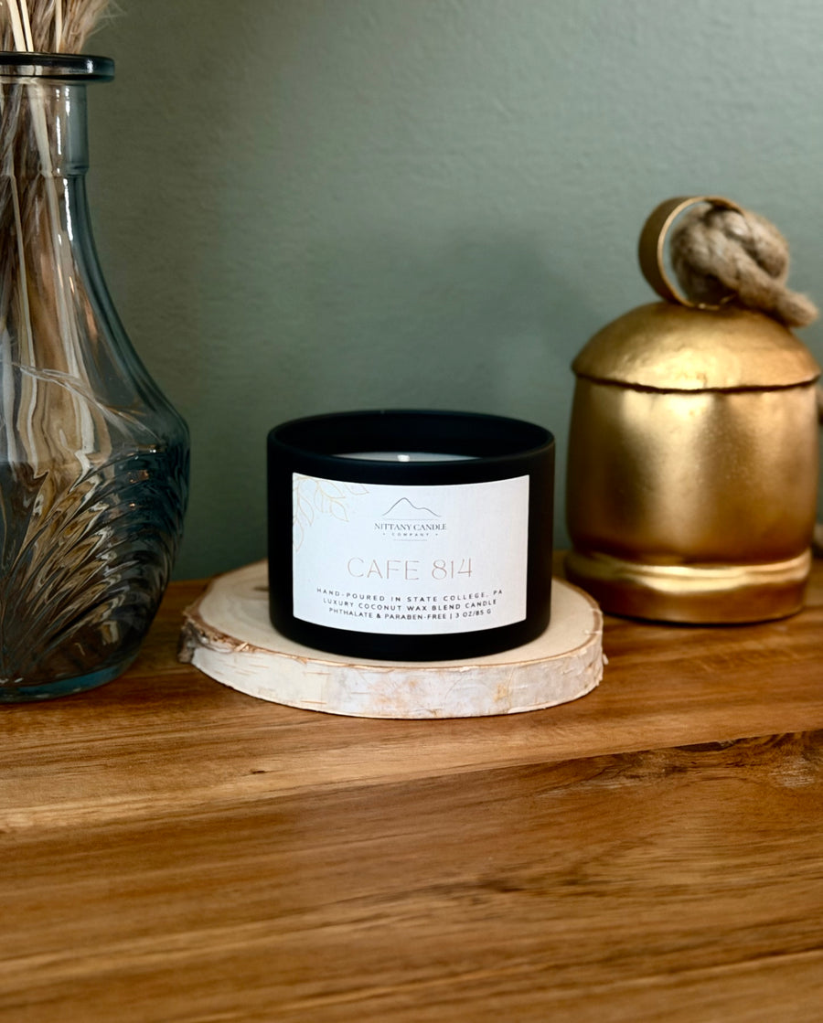 A black candle jar with a white label reading 'CAFE 814' sits on a round wood slice coaster on a wooden table, styled with a blue glass vase and a gold decorative container against a muted green wall.