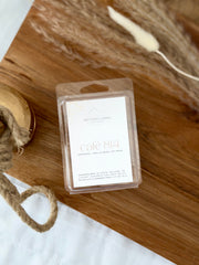 A packaged wax melt  labeled 'NITTANY CANDLE' and 'cafe 814' sits on a wooden board with decorative rope and dried grass. The product is shown in clear plastic packaging with a white label.