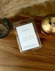 A clear plastic clamshell package containing a white wax melt label for 'Nittany Candle Company' scented with 'café 814', placed on a wooden surface next to a gold candle tin.