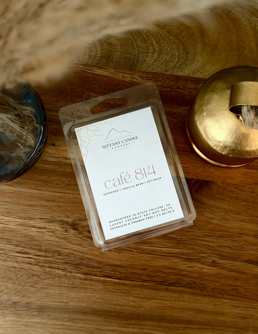 A clear plastic clamshell package containing a white wax melt label for 'Nittany Candle Company' scented with 'café 814', placed on a wooden surface next to a gold candle tin.