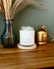 A jar candle labeled 'cider donut' sits on a wooden coaster on a wooden table, styled with a dark blue glass vase of dried grass on the left and a gold metal container on the right against a muted green wall.