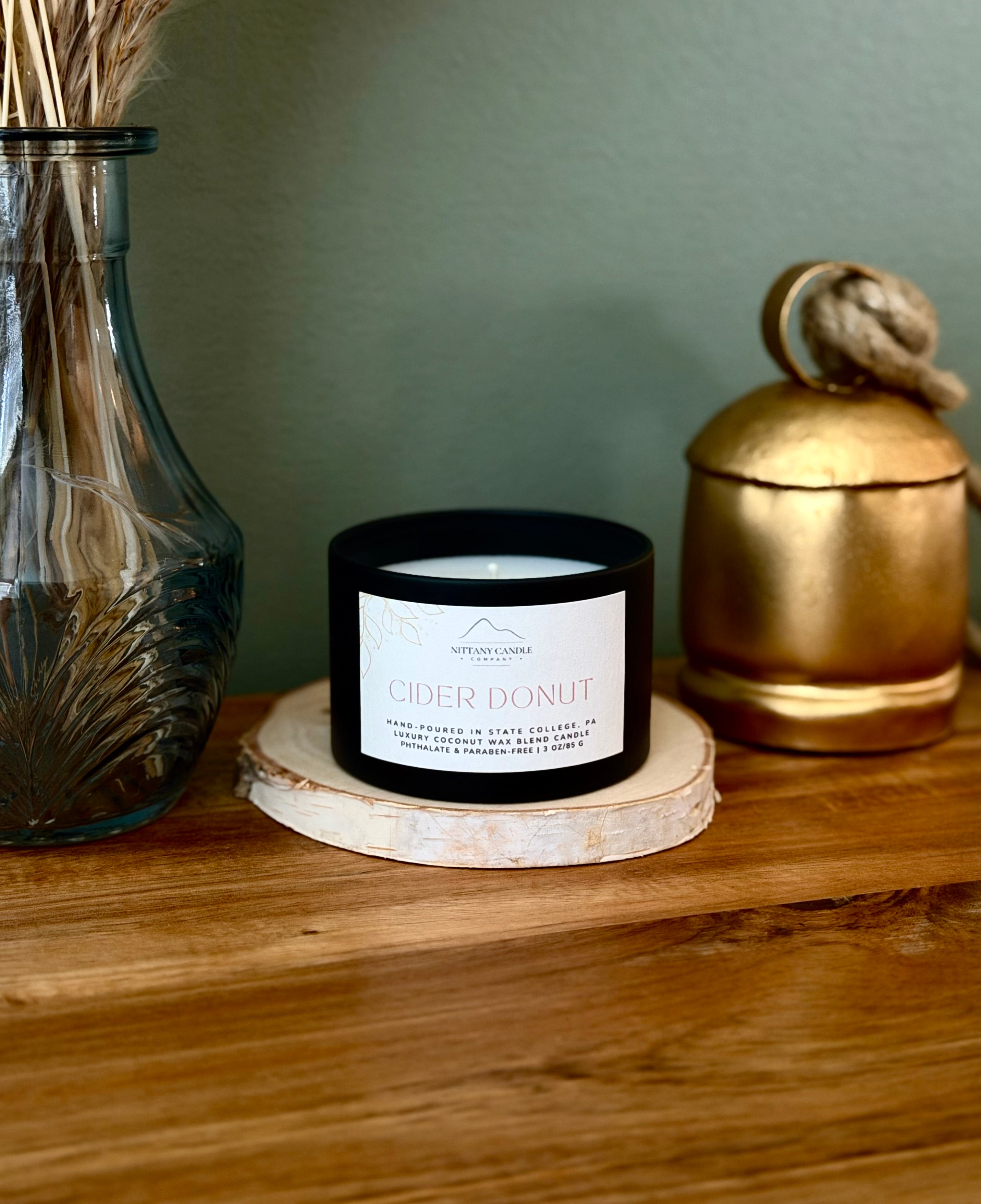 A black candle jar labeled 'CIDER DONUT' resting on a round wood slice coaster on a wooden table, styled with a blue glass vase and a gold container in the background.