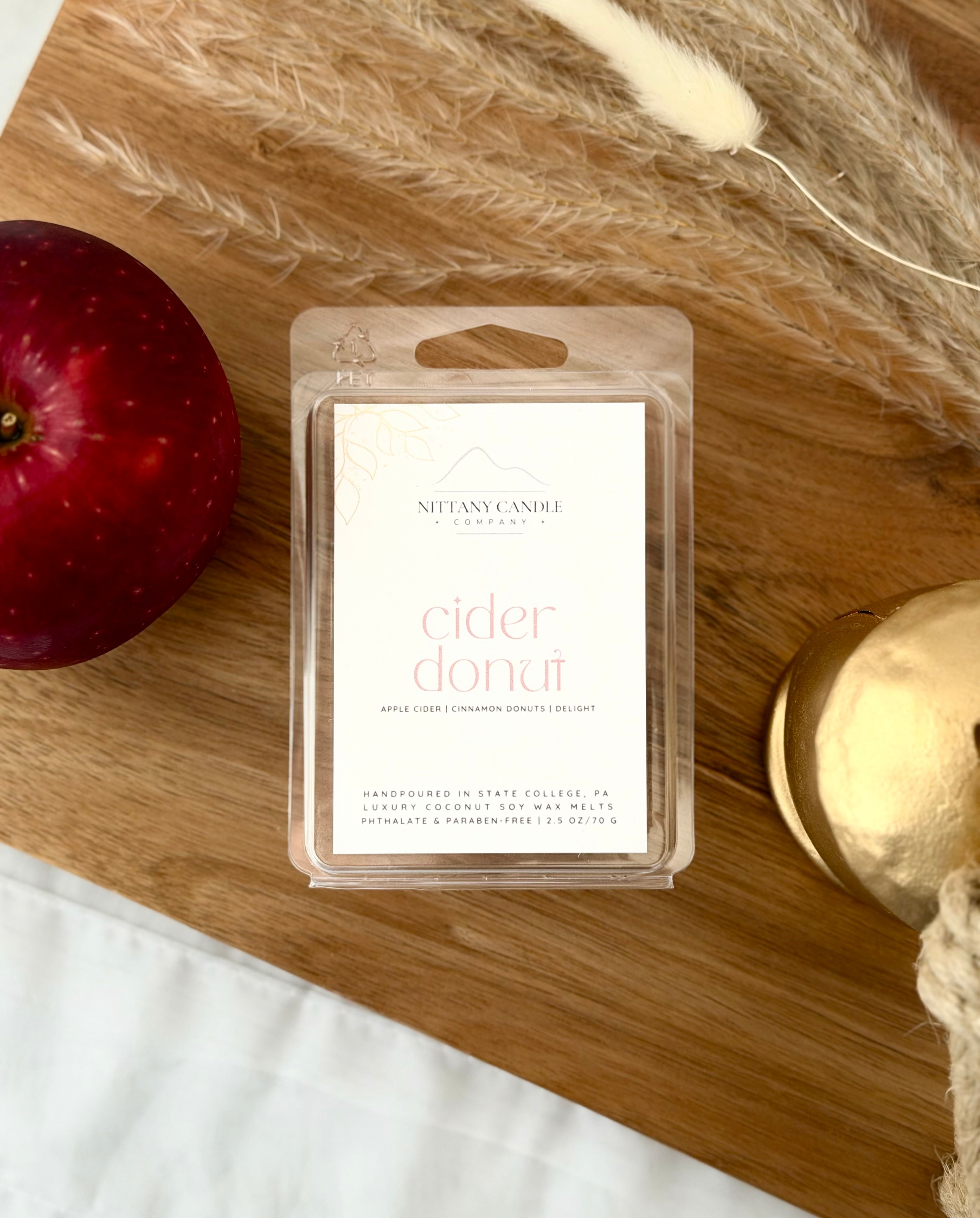 A clear plastic clamshell package containing a white wax melt label that reads 'NITTANY CANDLE COMPANY cider donut' in pink text, placed on a wooden board with a red apple, dried grass, and a gold container nearby.