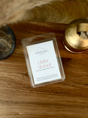 A clear plastic clamshell package of Nittany Candle Co. 'cider donut' scented wax melts sits on a warm wooden tabletop, styled with a gold-toned container and a dark glass vessel in the background.