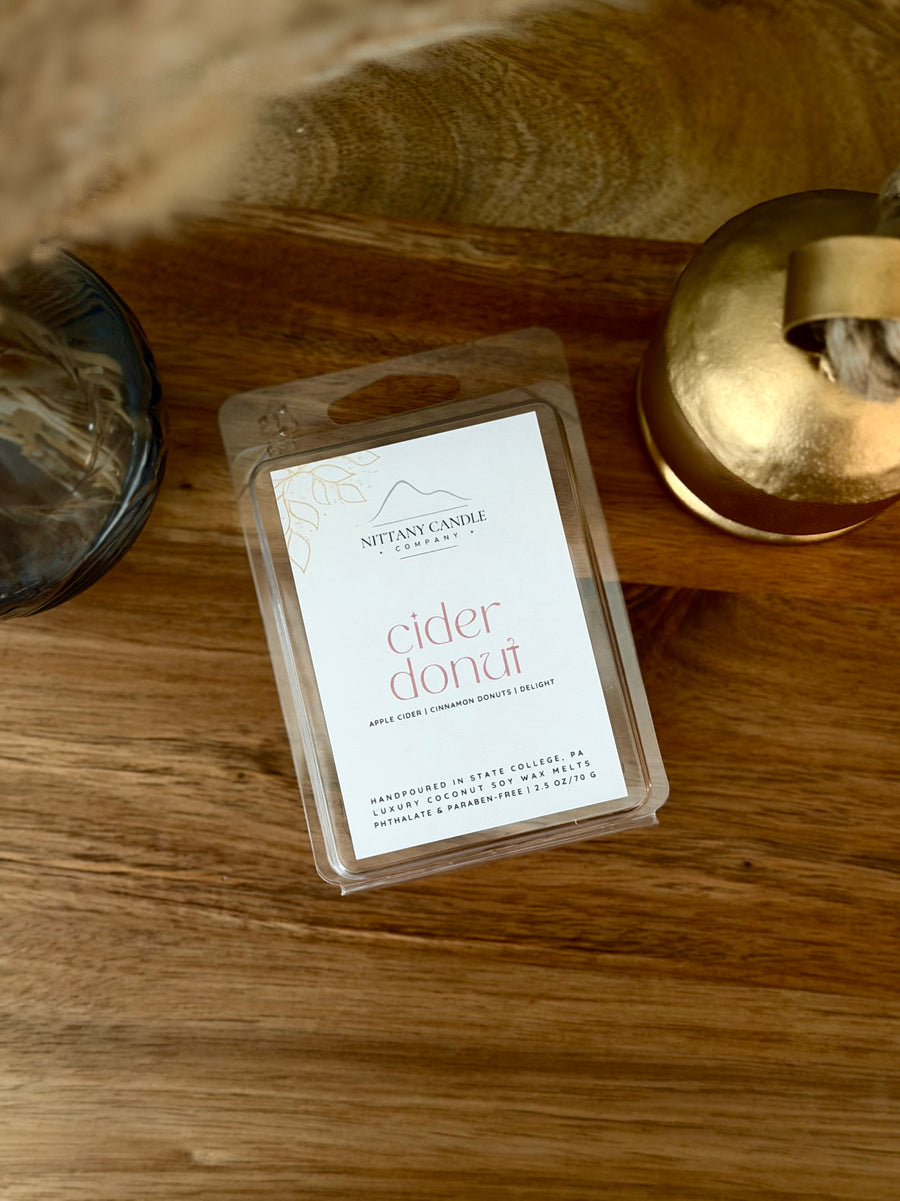 A clear plastic clamshell package of Nittany Candle Co. 'cider donut' scented wax melts sits on a warm wooden tabletop, styled with a gold-toned container and a dark glass vessel in the background.