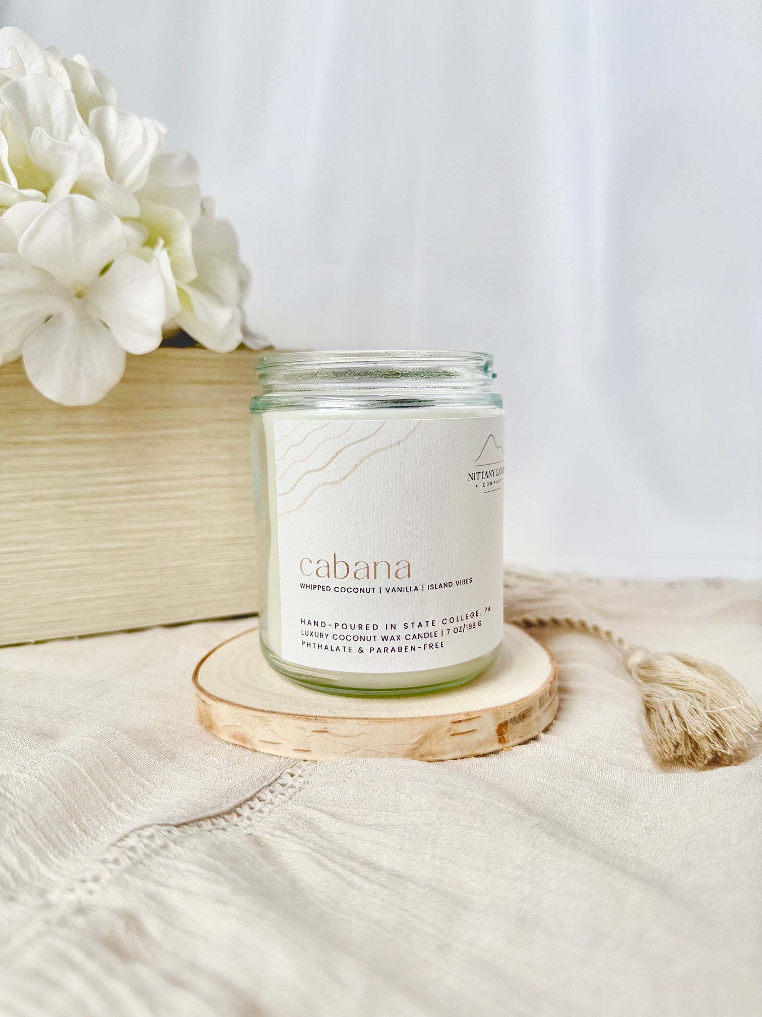 Candle labeled 'acabana' on a wooden coaster with white flowers in the background