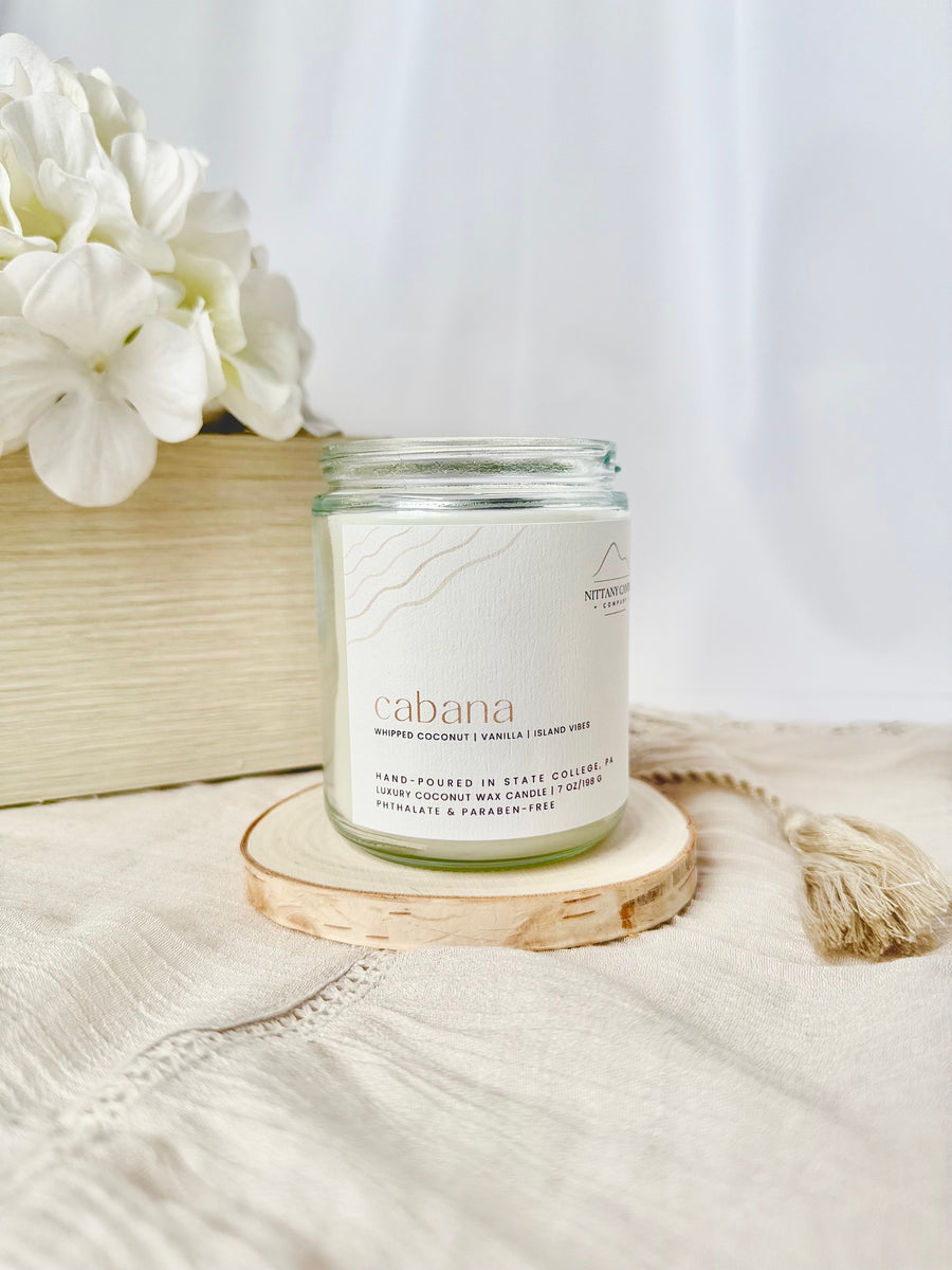 Candle labeled 'acabana' on a wooden coaster with white flowers in the background