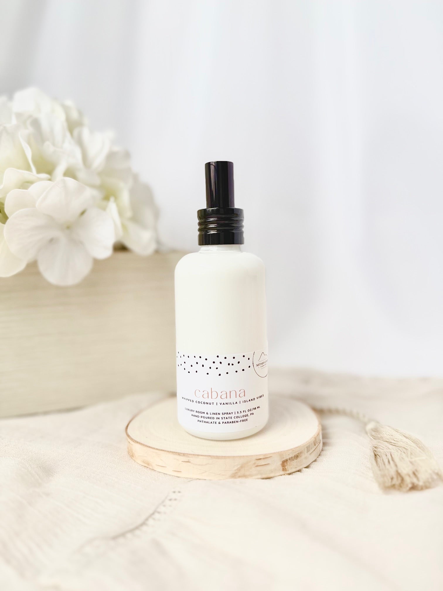 White bottle with black cap on a wooden coaster, surrounded by white flowers and fabric.