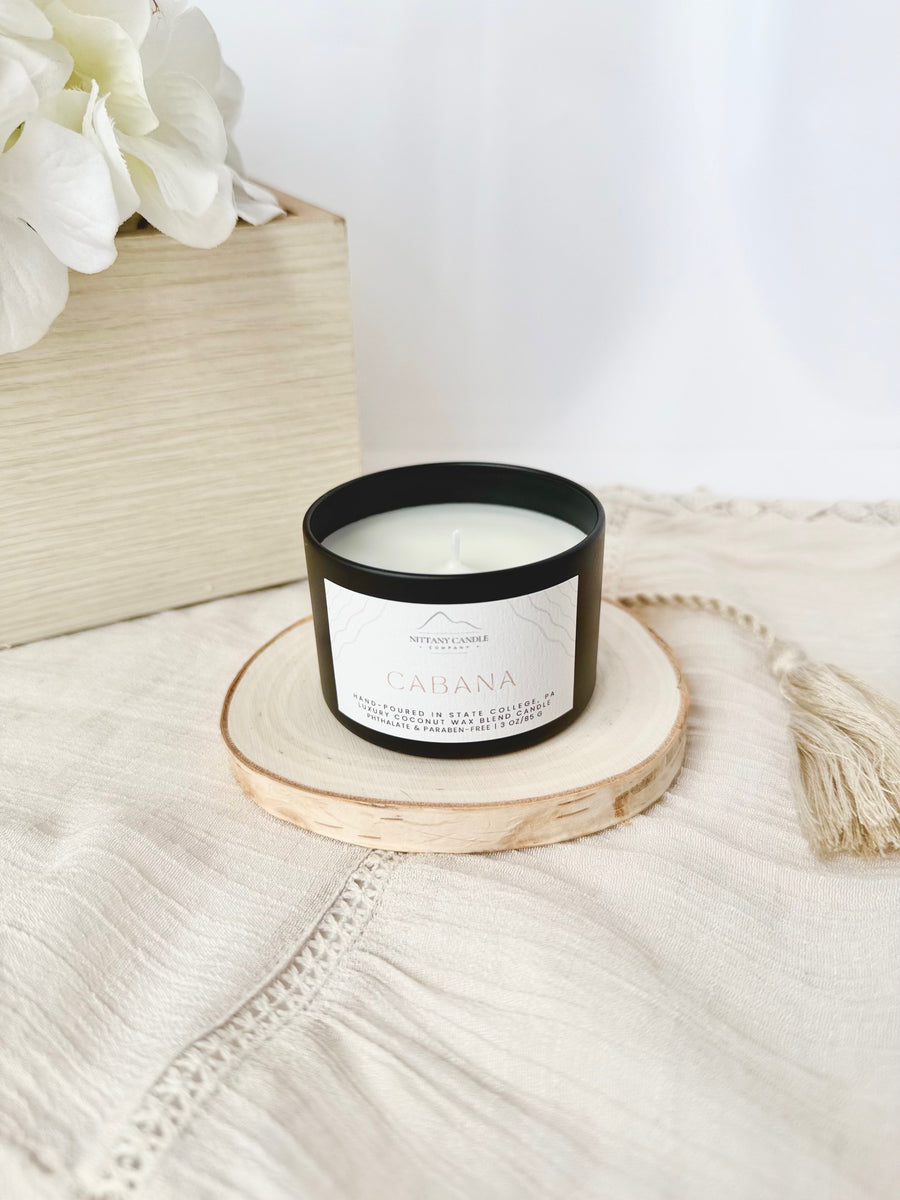 Candle in a black container and white label on a wooden coaster, surrounded by white flowers and a beige woven basket.