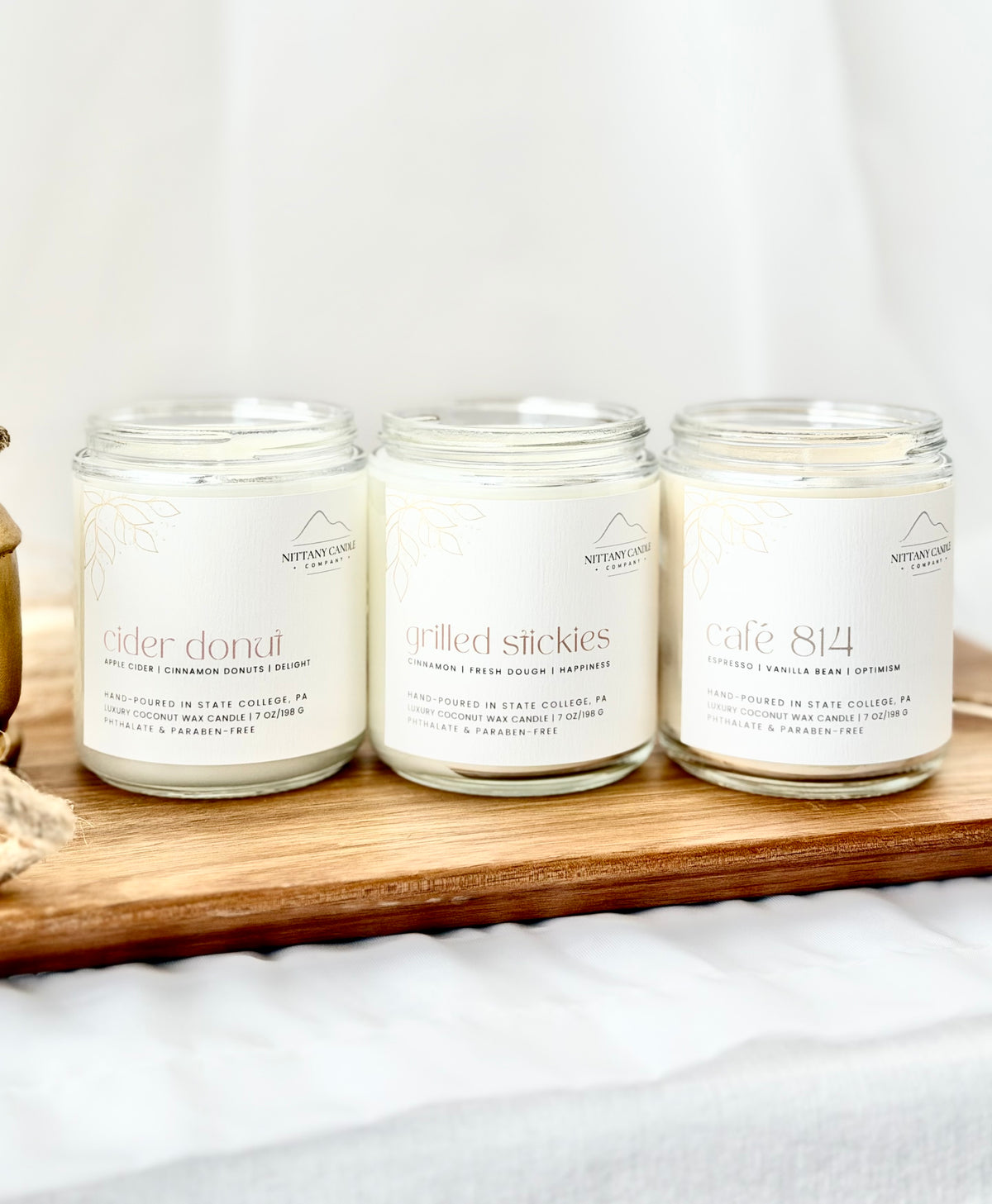 Three natural soy candles in clear glass jars with white labels, arranged on a wooden tray. The labels read 'Nature's Way' and show scents 'cider donut', 'grilled stickles', and 'cafe 814'.