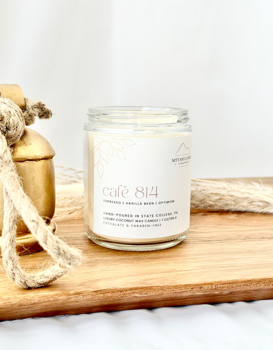 A glass jar candle labeled 'cafe 814' sits on a wooden board with rustic twine and a gold-toned decorative object in the background. The candle has a white label with elegant text and a small mountain logo.