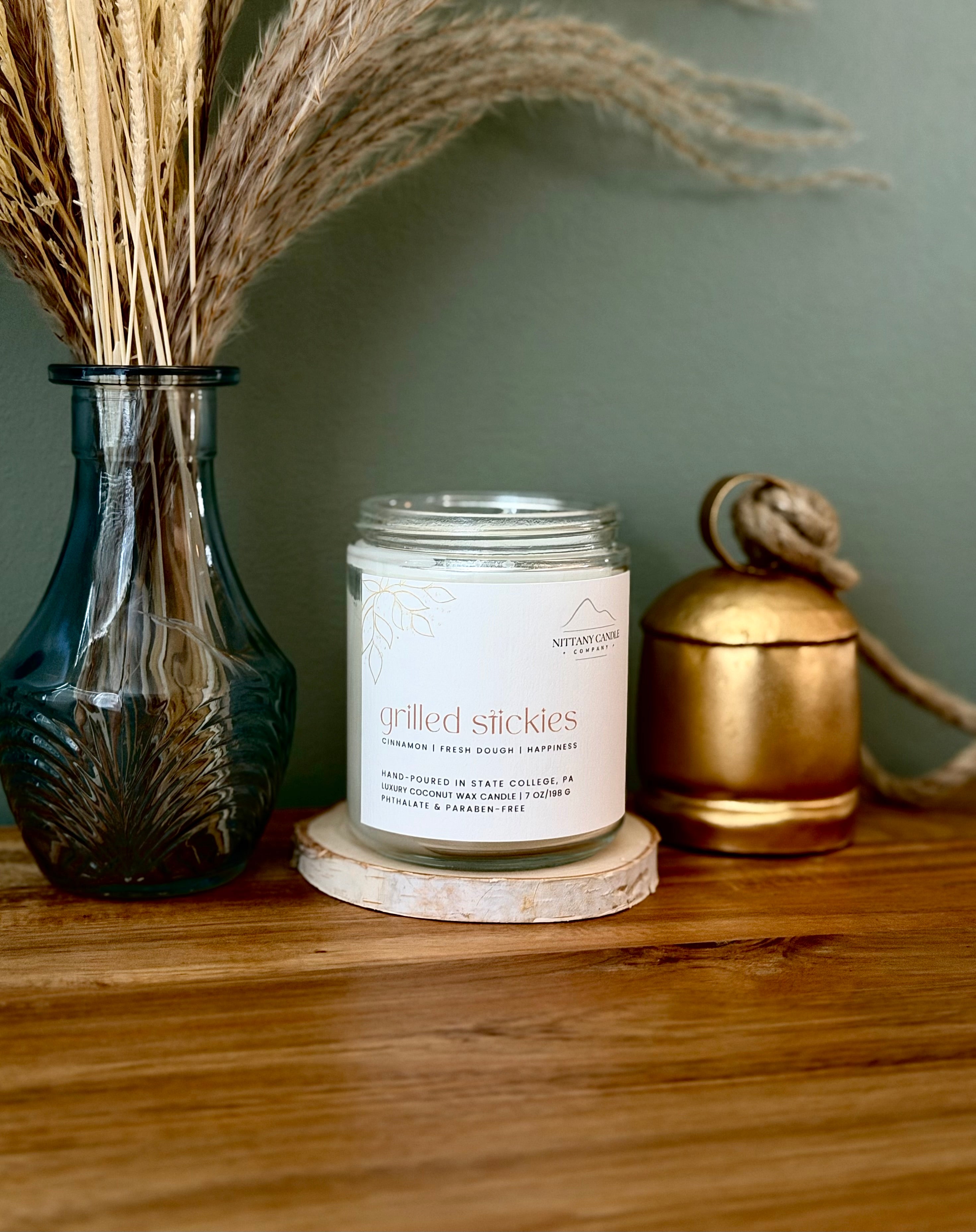 A glass jar candle labeled 'grilled stickies' sits on a wooden coaster on a wooden table, styled with a blue glass vase of dried grass on the left and a gold metal container on the right against a muted green wall.