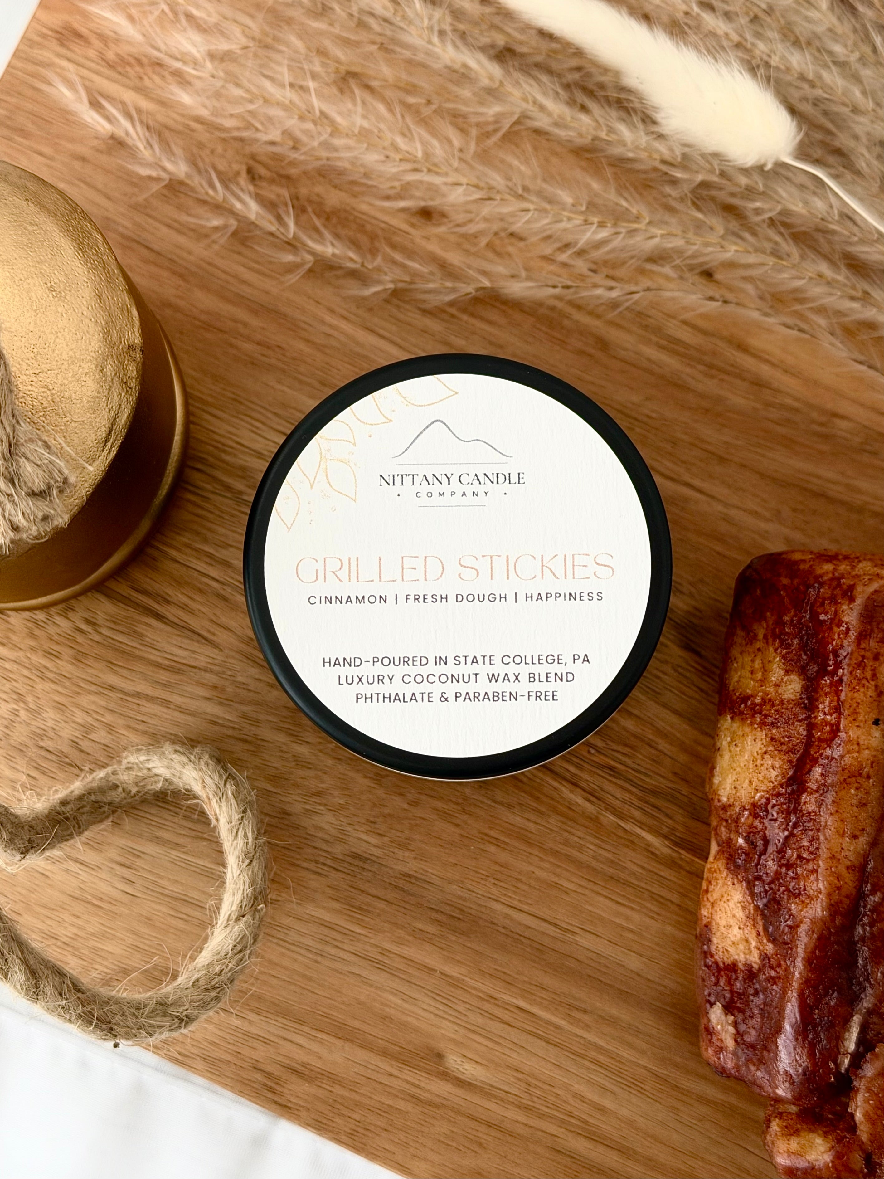 A round tin candle of Nittany Candle's 'Grilled Stickies' scent sits on a wooden board styled with dried grass, rope, a gold bell, and sliced of cinnamon roll. The label is white with black and gold text.