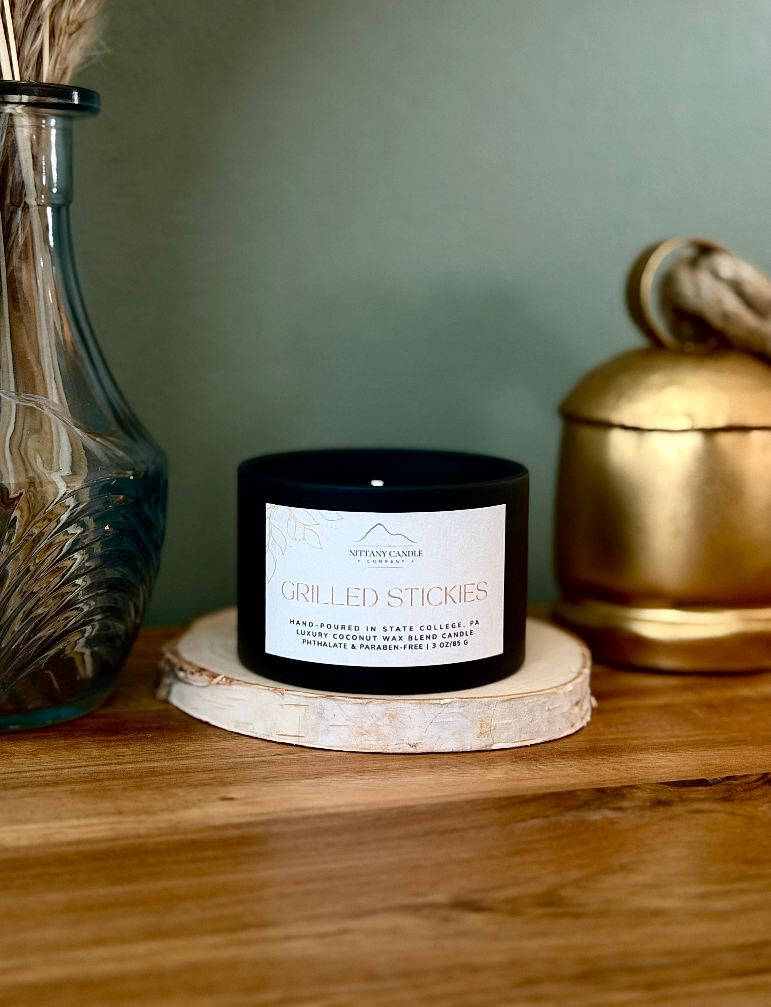 A black candle jar labeled 'Grilled Stickies' by Nittany Candle Co., styled on a wooden slice coaster on a wooden table with a blue glass vase and a gold decorative container in the background.