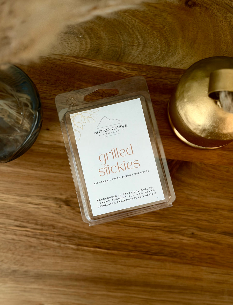 A clear plastic clamshell package of Nittany Candle Company 'grilled stickies' wax melts sits on a rustic wooden surface, flanked by a gold candle tin and a dark glass container.