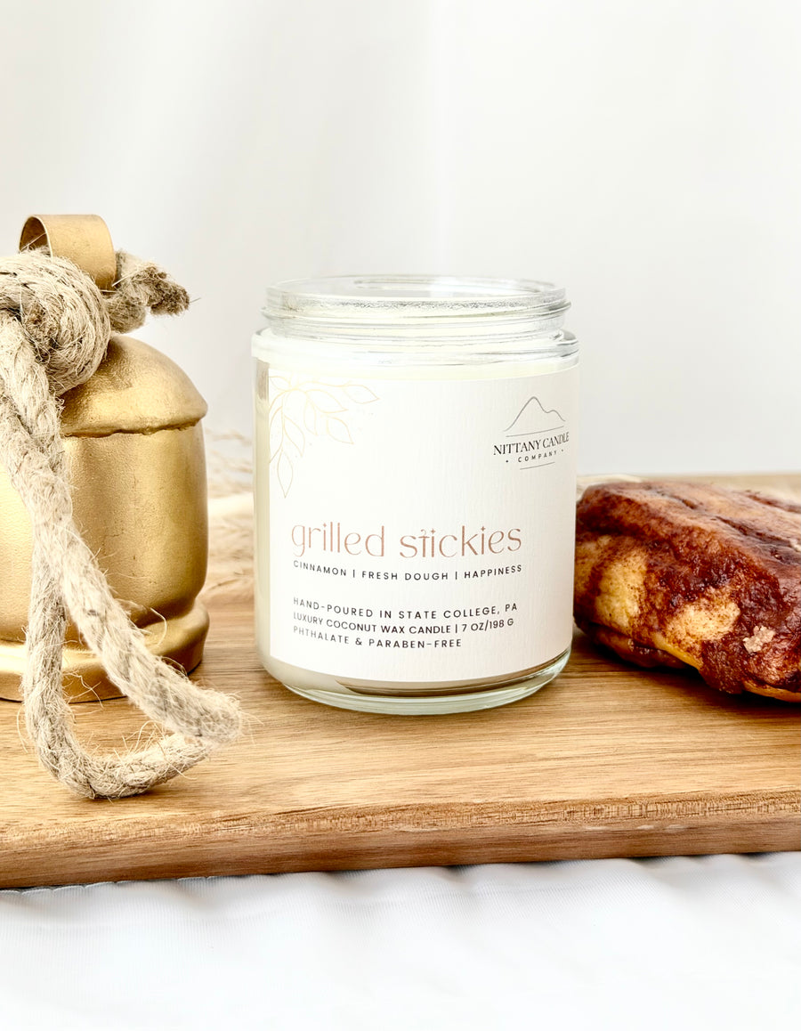 A glass jar candle labeled 'grilled stickies' by Nittany Candle Co. sits on a wooden board, styled with a gold bell and rope on the left and a piece of chocolate cake on the right.