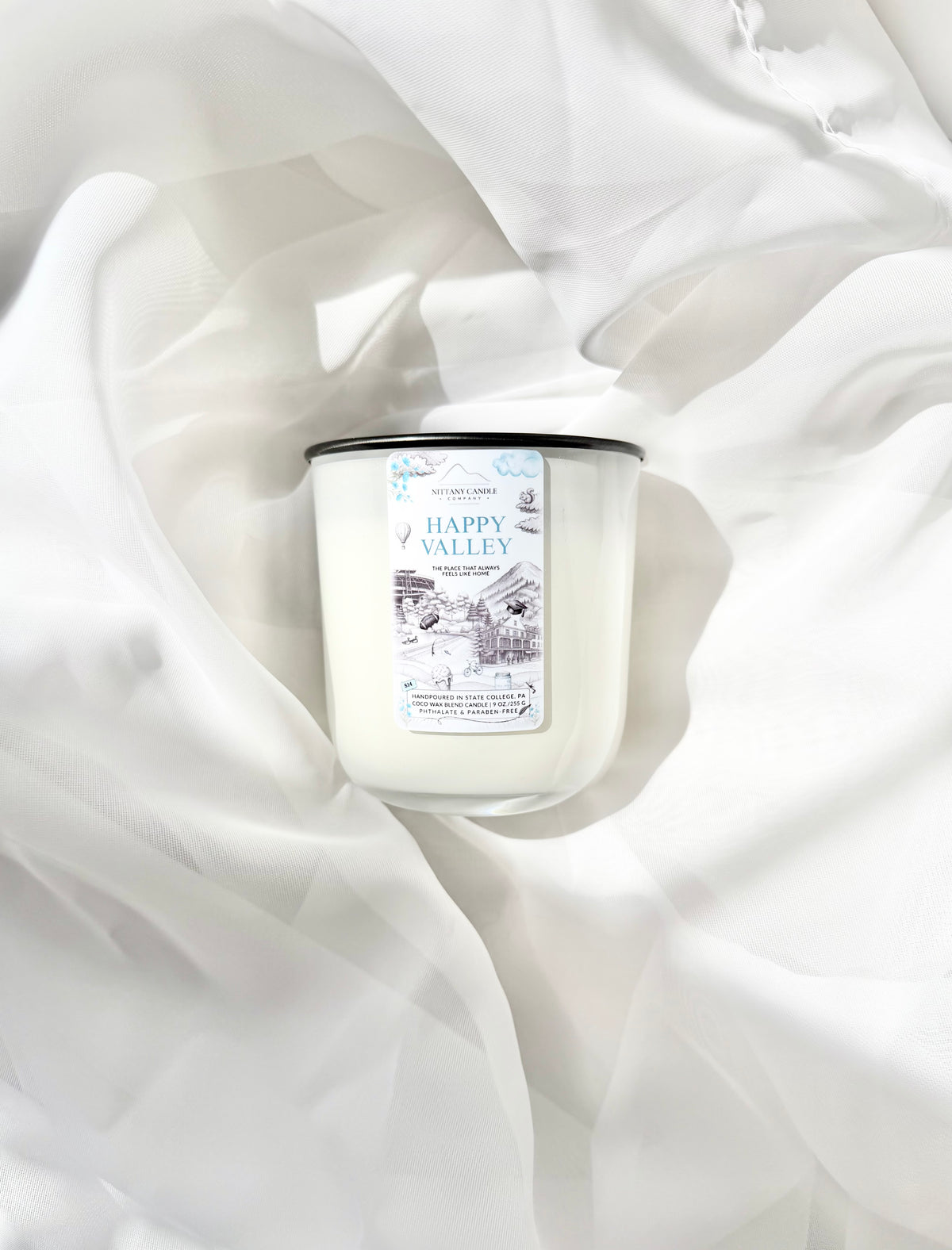 A white scented candle with a black lid and a white label reading 'Happy Valley,' styled on draped white fabric.
