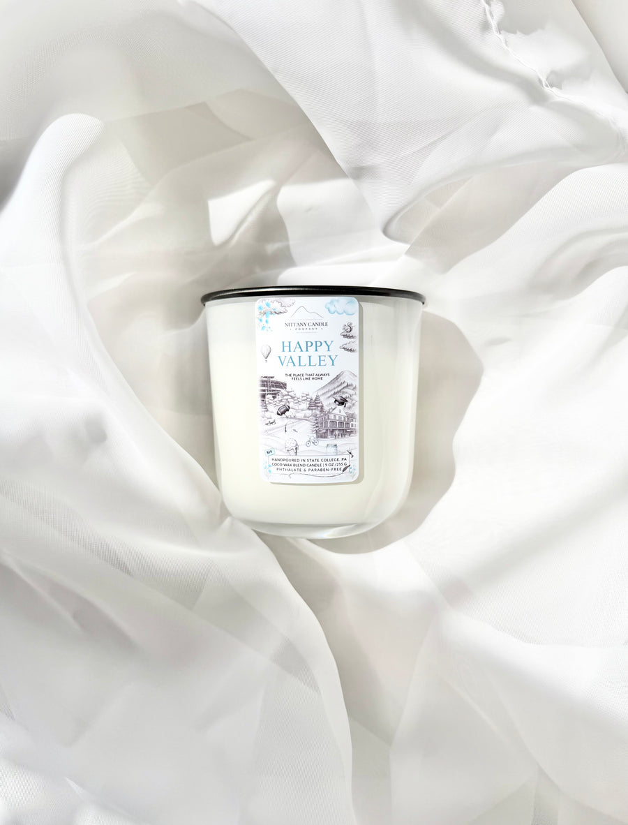 A white scented candle with a black lid and a white label reading 'Happy Valley,' styled on draped white fabric.