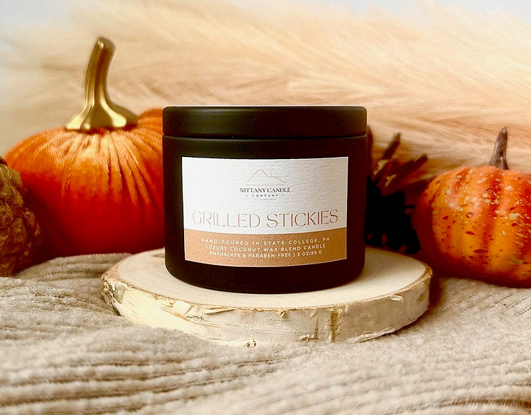 Nittany Candle Company candle labeled 'Grilled Stickies' on a wooden surface with pumpkins in the background