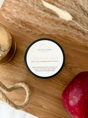 A round tin candle labeled 'Nittany Candle Company Cider Donut' sits on a wooden board with decorative dried grass, rope, a gold bell, and a red apple nearby.