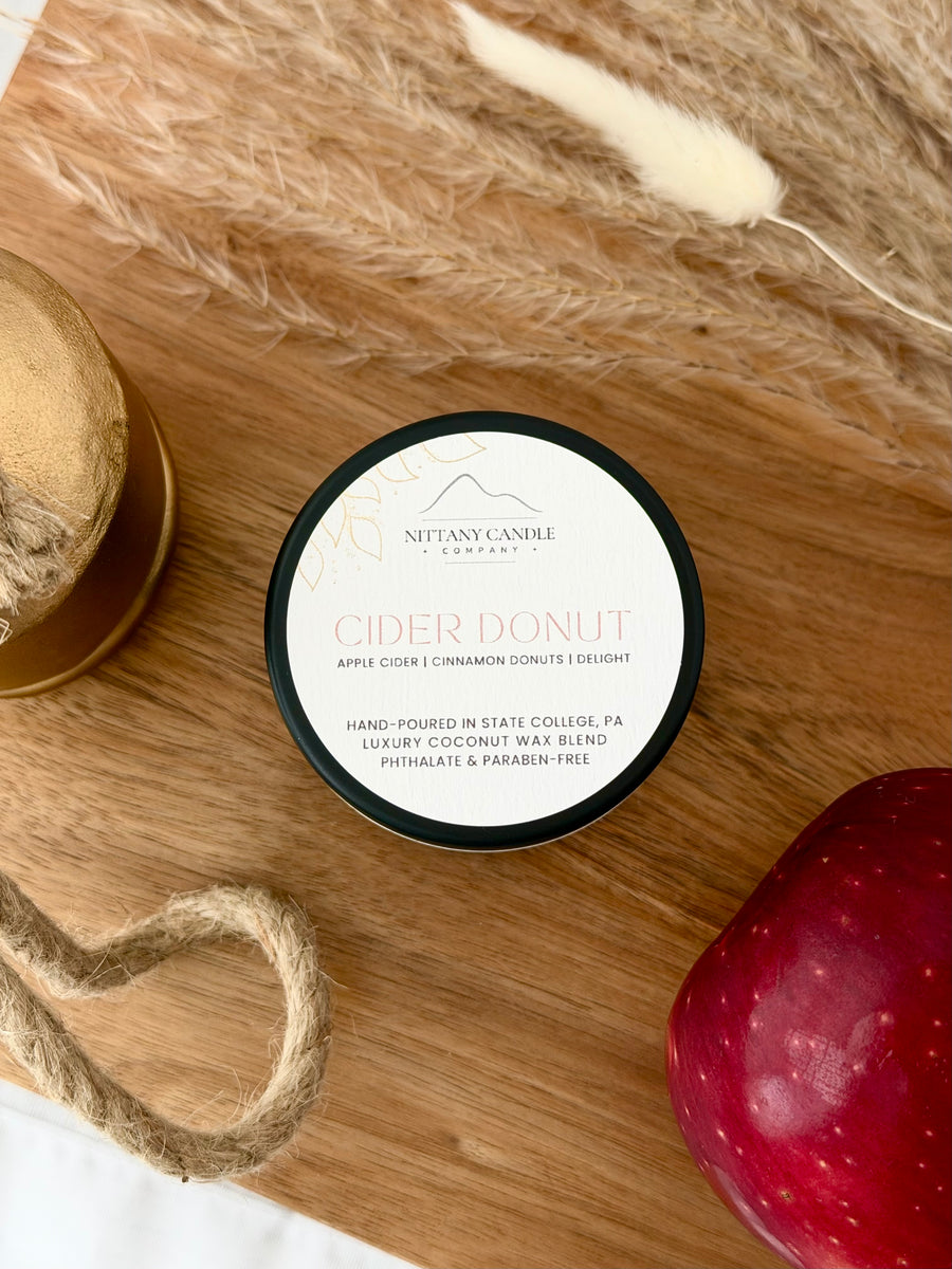 A round tin candle labeled 'Nittany Candle Company Cider Donut' sits on a wooden board with decorative dried grass, rope, a gold bell, and a red apple nearby.