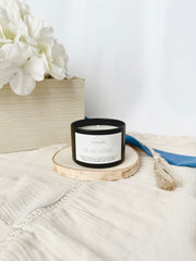 Black candle with a white label on a wooden coaster, surrounded by white flowers and a blue towel on a light surface.