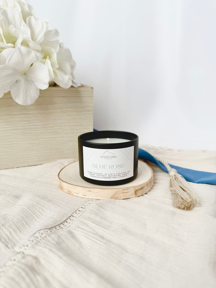 Black candle with a white label on a wooden coaster, surrounded by white flowers and a blue towel on a light surface.