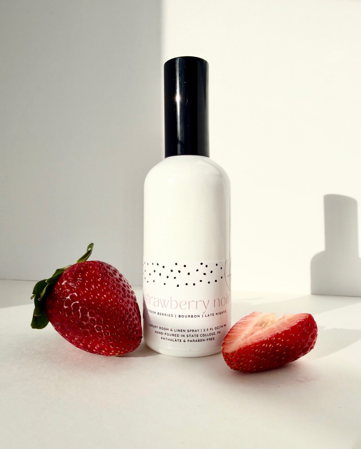 Nittany Candle Company linen spray in a white bottle with black cap on a white surface with strawberries around it