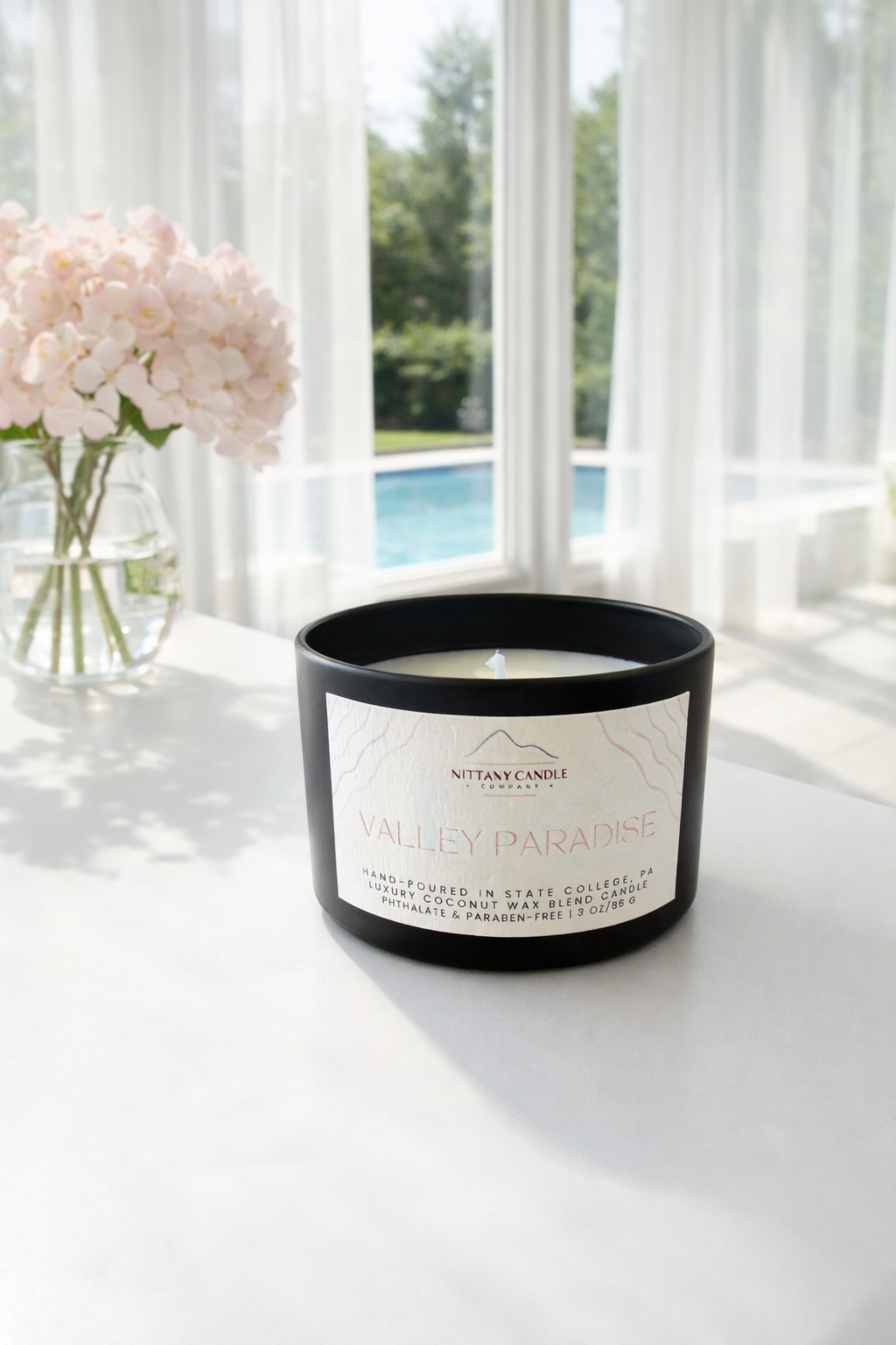 Small Nittany Candle Company candle on a white table with pink flowers and a pool in the backgrond