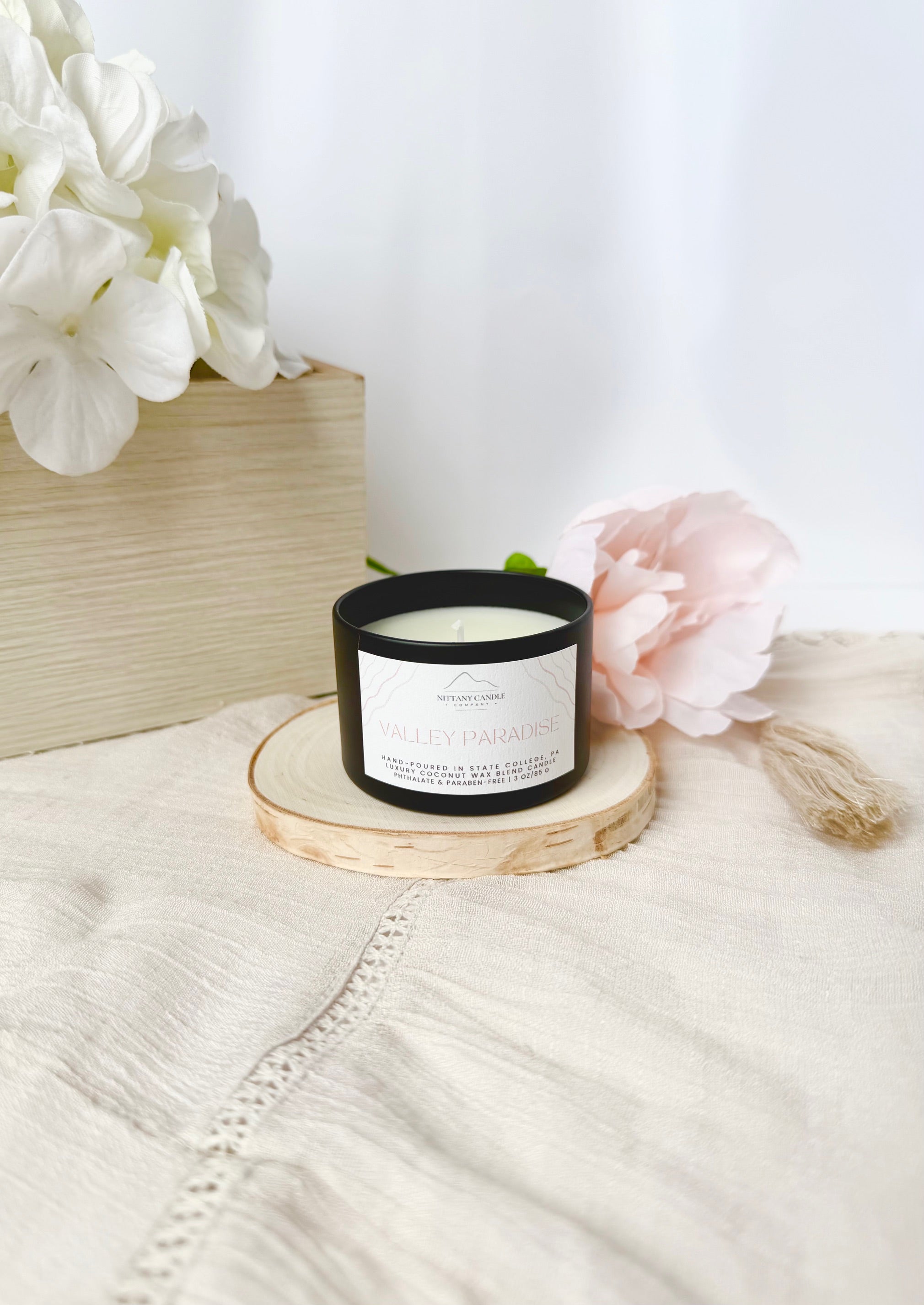 Candle with a label on a wooden coaster surrounded by flowers on a light surface.
