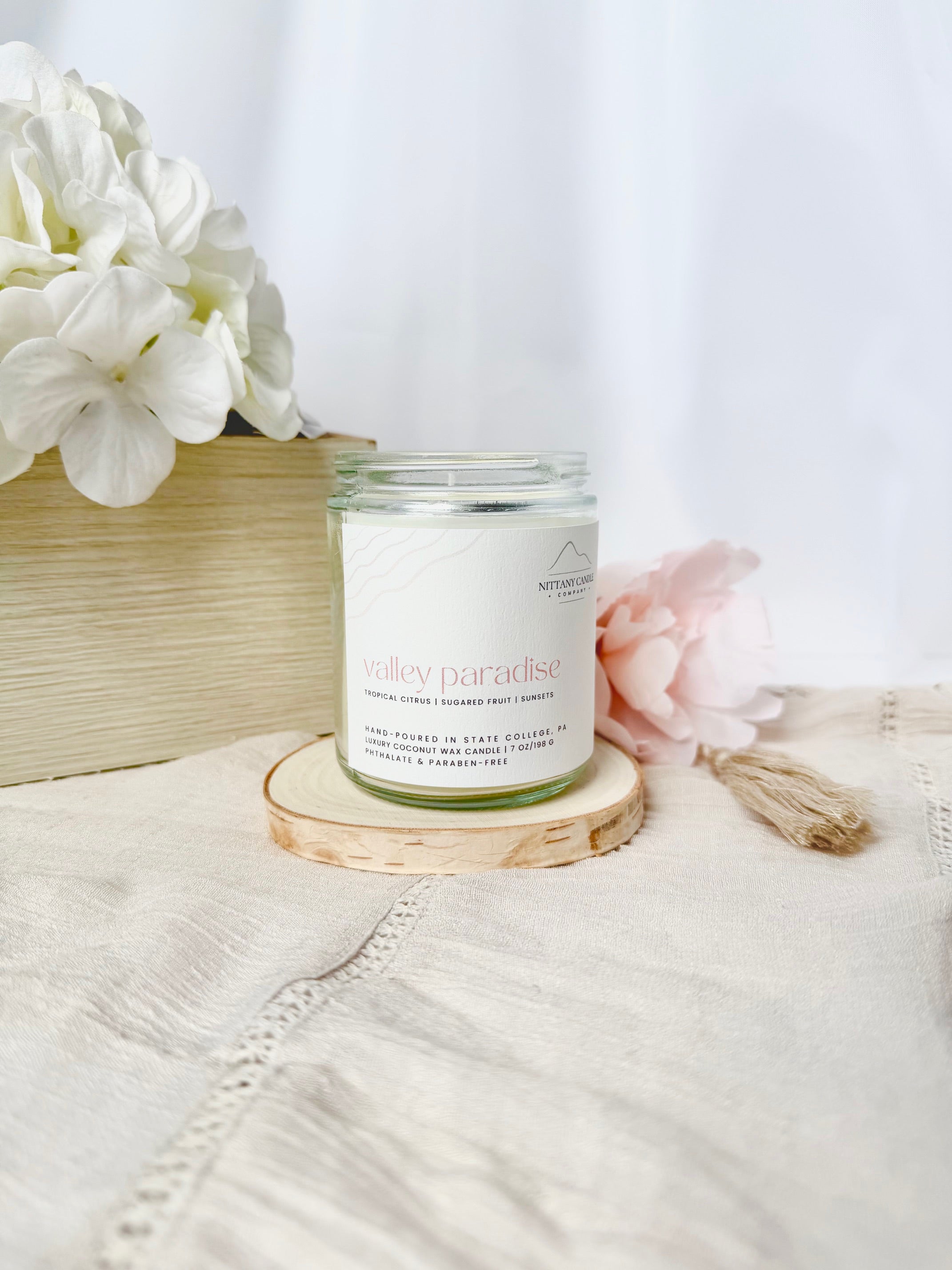 Candle labeled 'Paradise' on a wooden coaster with flowers in the background